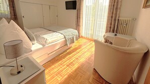 A hotel room with a bed, a chair, a desk with a lamp, a TV on the wall, and a window with curtains. - Apartment, 55sqm with 8sqm balcony, 2-4persons in Baiersbronn center (Baiersbronn)
