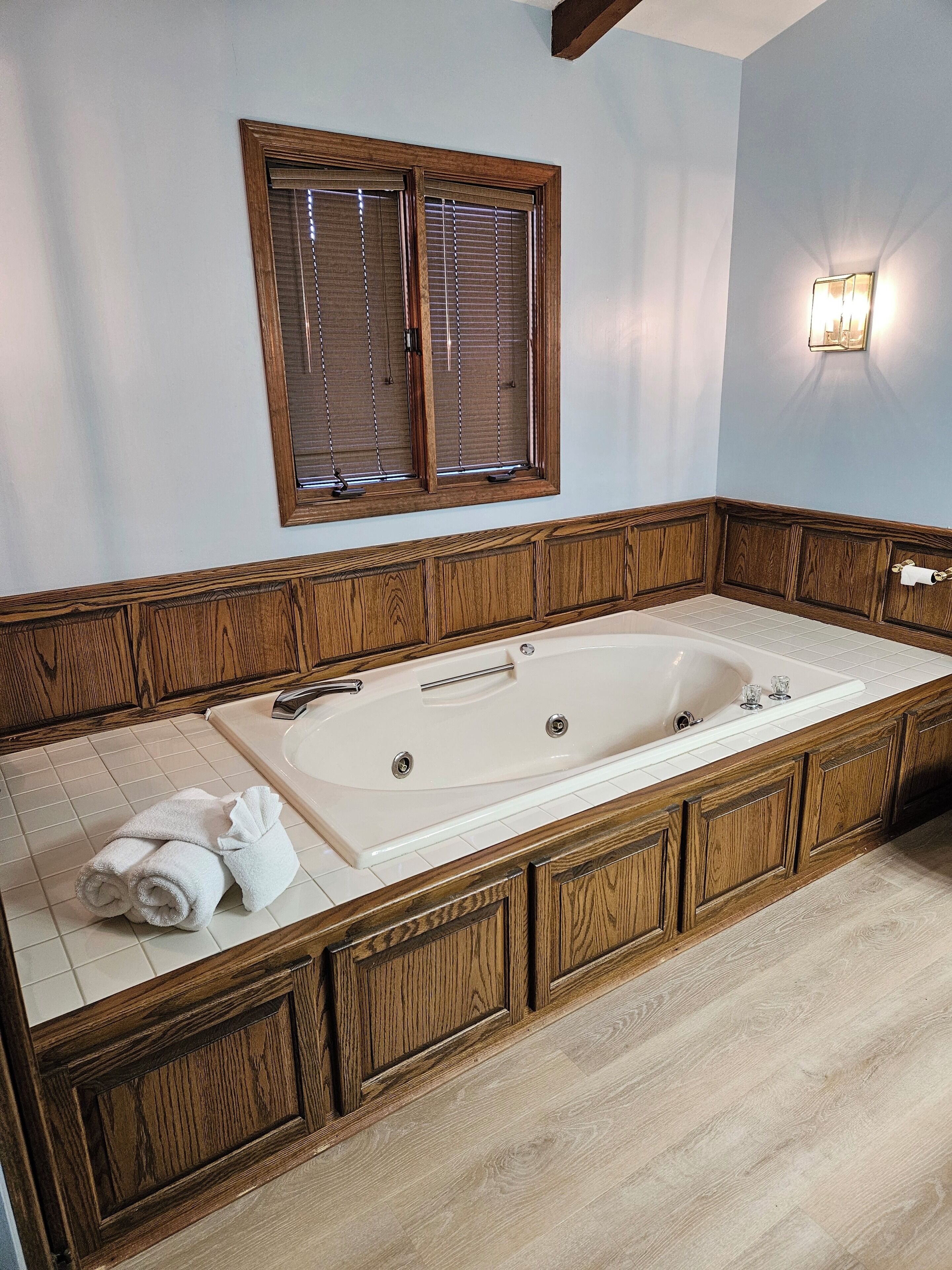 A bathroom with a large bathtub, wooden cabinetry, a window with blinds, and a wall-mounted light fixture.