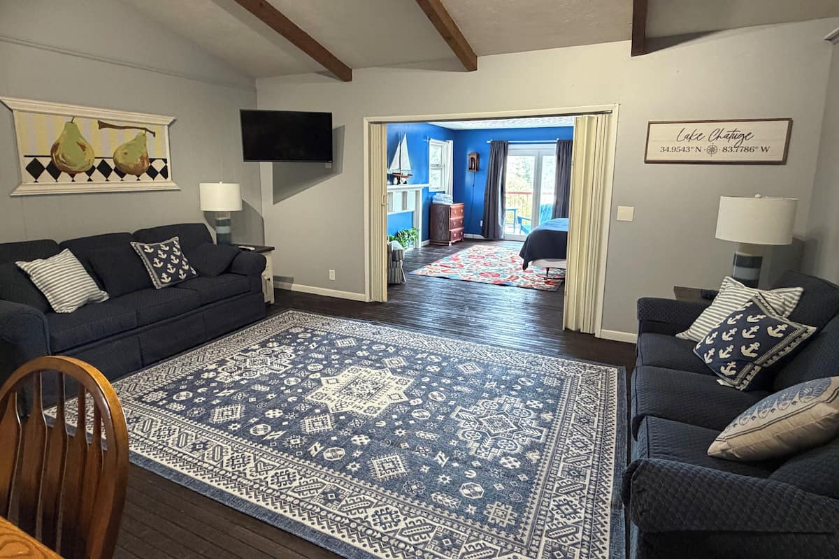 A living room with a dark blue sofa, a matching armchair, a patterned rug, a television, and a door leading to a balcony.