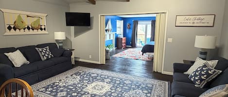 A living room with a dark blue sofa, a matching armchair, a patterned rug, a television, and a door leading to a balcony.