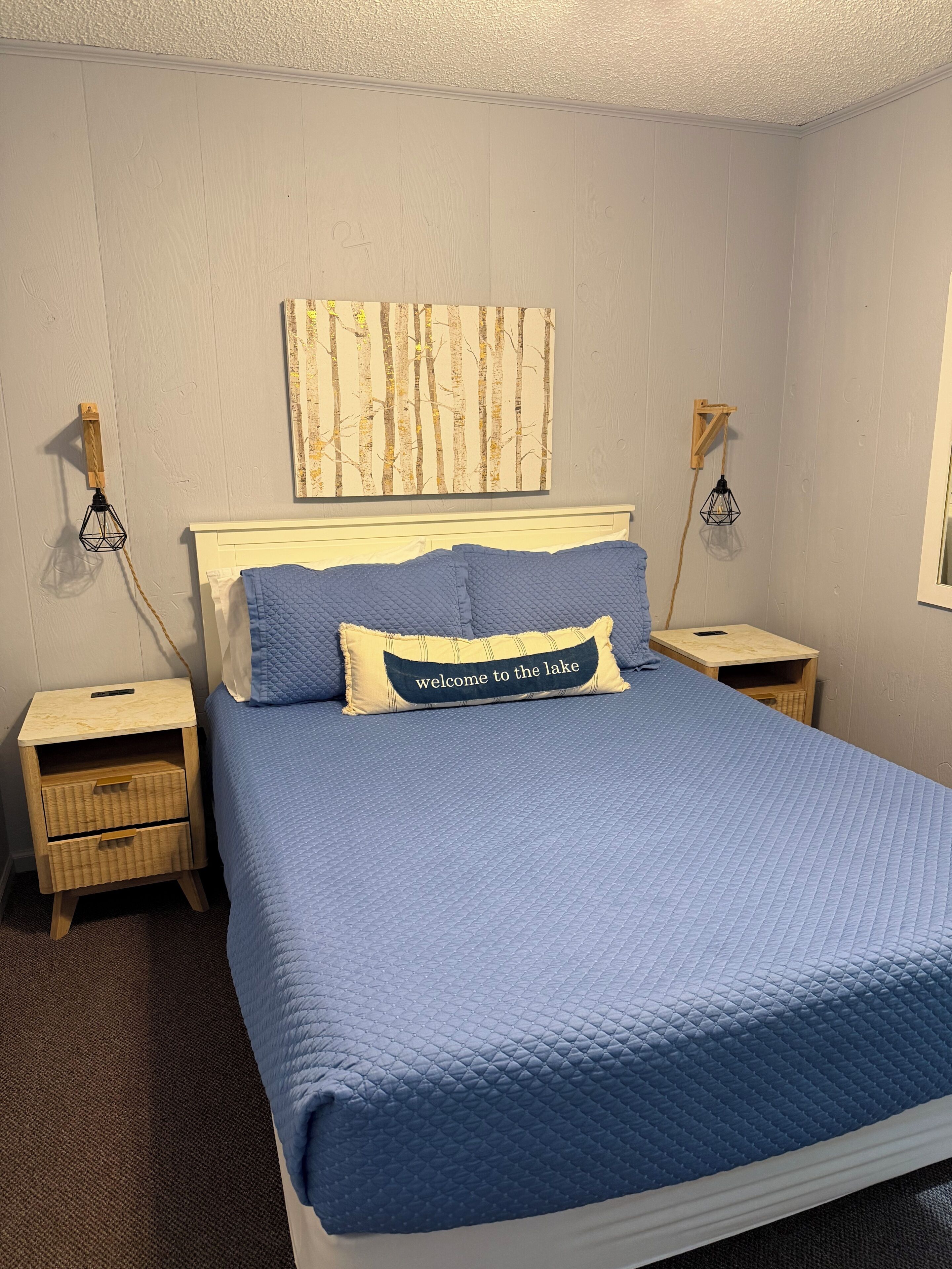 A bedroom with a bed, two bedside tables, a nightstand, a wall-mounted lamp, and a framed picture.