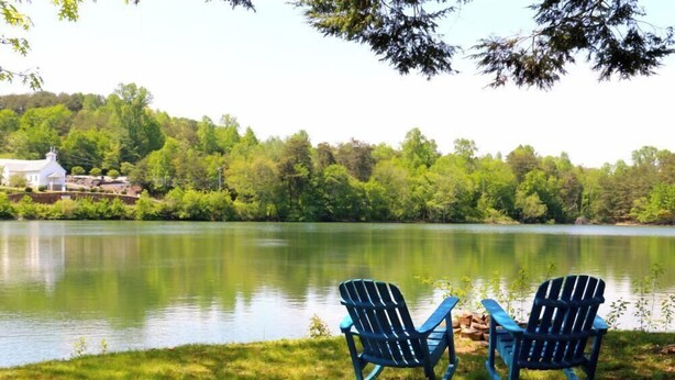 Två Adirondack-stolar vid en sjö, med en vit byggnad i bakgrunden.