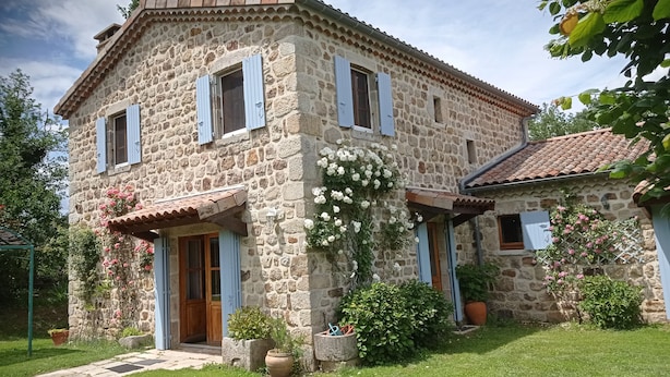 Une maison en pierre avec une porte en bois, des volets bleus et un jardin agrémenté de plantes et de fleurs en pot.