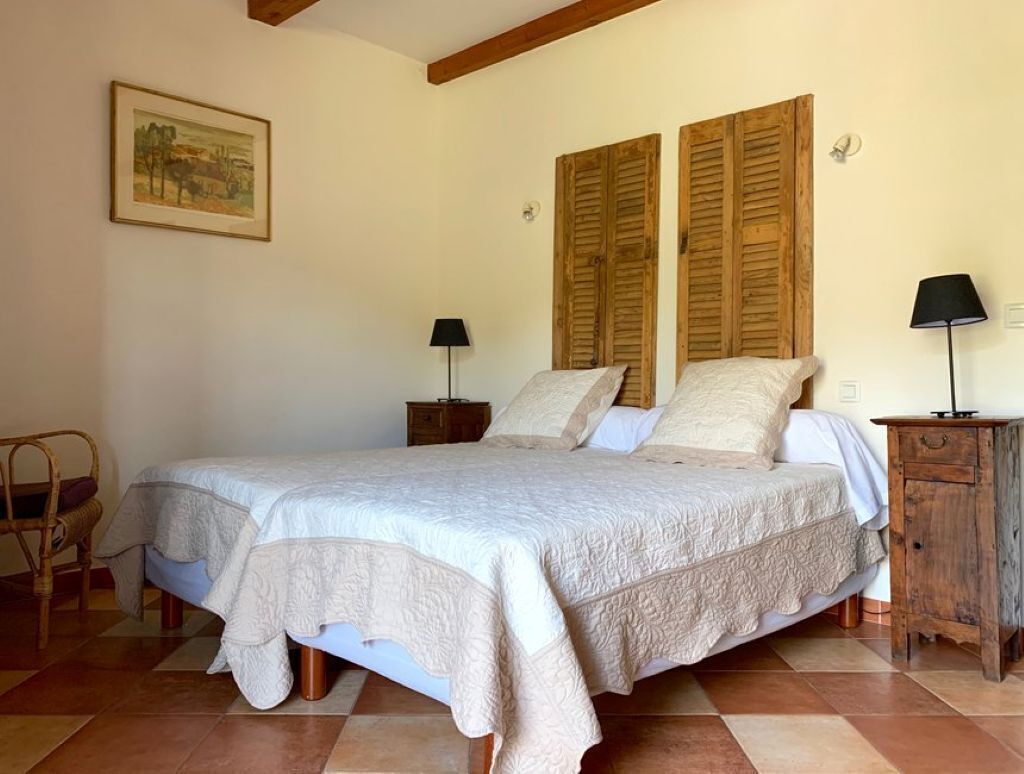 Een slaapkamer met een groot bed, houten luiken, twee nachtkastjes, een stoel en een lamp.