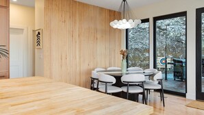 A modern dining area with a wooden table and white chairs, a wooden slatted wall, and a large window with a view of a snowy landscape. - Ava A-Frame•Hot Tub•Theater•Hidden Stair•EV Charge (Stanley)
