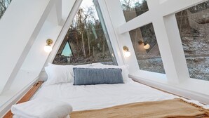 A bedroom with a bed, pillows, and a blanket. There are windows with lights and a view of trees and snow outside. - Ava A-Frame•Hot Tub•Theater•Hidden Stair•EV Charge (Stanley)