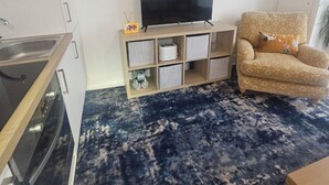 A living room with a TV on a wooden stand, a sofa, and a kitchen area with cabinets and a microwave. - Seaside Haven Coastal Comfort 200m from the Beach (Margate)