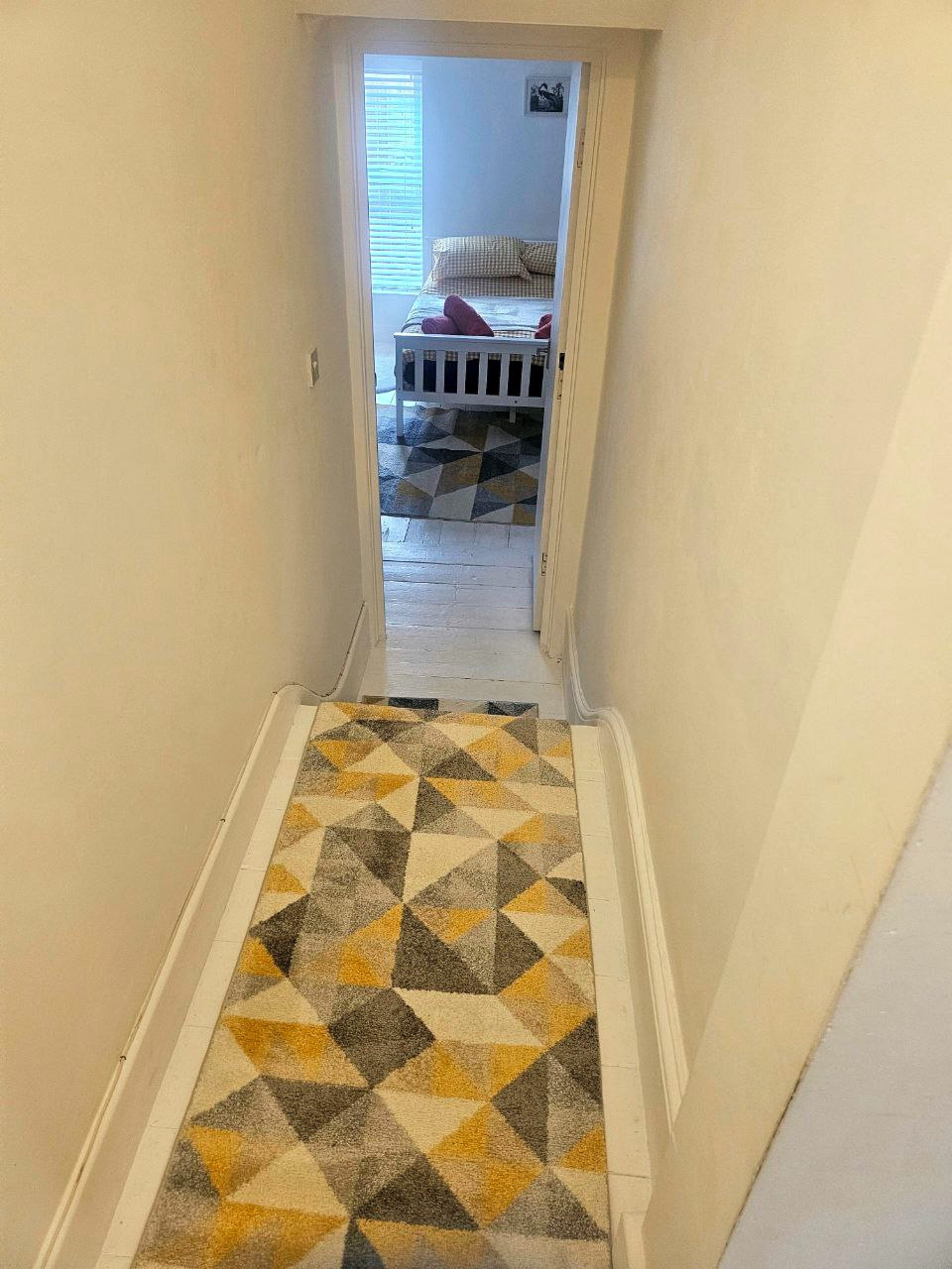 A narrow hallway with a geometric-patterned carpet leading to a bedroom with a bed and a checkered pillow.