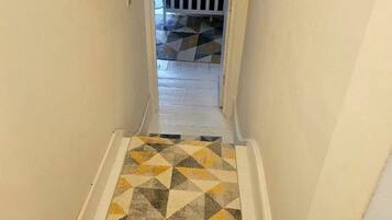 A narrow hallway with a geometric-patterned carpet leading to a bedroom with a bed and a checkered pillow.