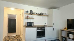 A compact kitchen with white cabinets, a microwave, a stove, and a sink. There are wooden shelves above the counter holding dishes and jars. A television is on the right side of the room. - Seaside Haven Coastal Comfort 200m from the Beach (Margate)