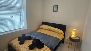 A bedroom with a bed, a bedside table with a lamp, a window with blinds, and a small picture on the wall. - Seaside Haven Coastal Comfort 200m from the Beach (Margate)