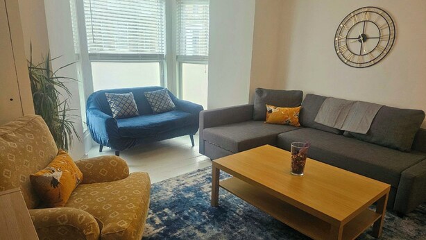 A living room with a sofa, armchair, and coffee table, a clock on the wall, and a window with blinds. - Seaside Haven Coastal Comfort 200m from the Beach (Margate)