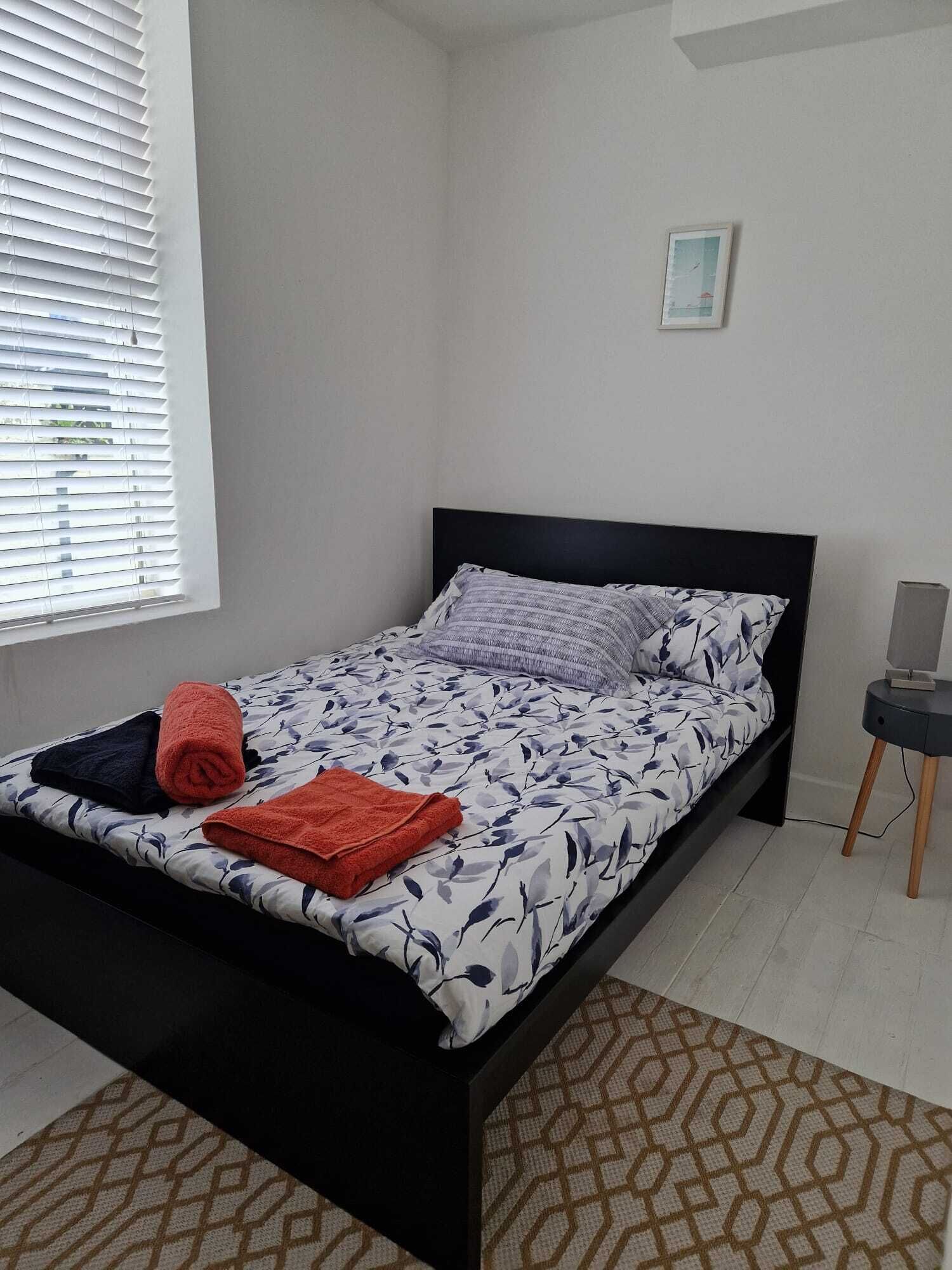 Une chambre à coucher avec un lit, une fenêtre avec des stores, une petite table avec un haut-parleur et un tapis à motifs.