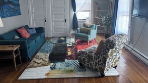 A living room with a teal sofa, a patterned armchair, a glass coffee table, and a colorful rug. There is a painting on the wall, a television, and a potted plant.