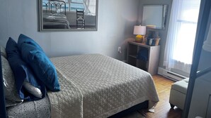 A bedroom with a bed, a chair, a bedside table with a lamp, a mirror, and a window with curtains.