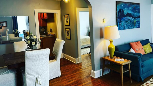 A living room with a blue sofa, a wooden coffee table, a white armchair, and a dining area with a mirror and flowers.