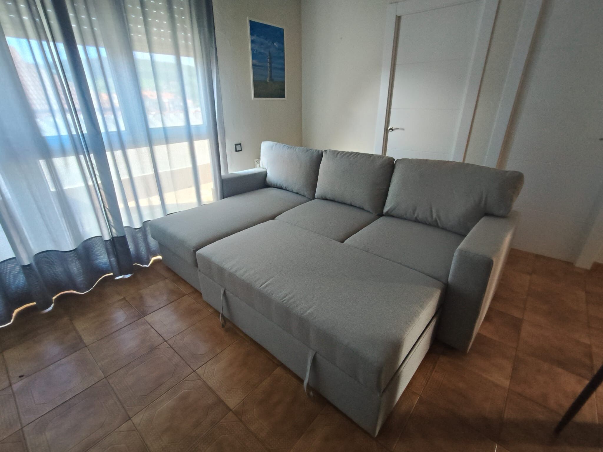 Un sofá cama gris en una habitación con suelo de baldosas y una ventana con cortinas ligeras.