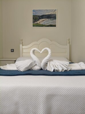 A bed with a white headboard, a painting of a coastal landscape above it, and a heart-shaped towel arrangement on the bed.