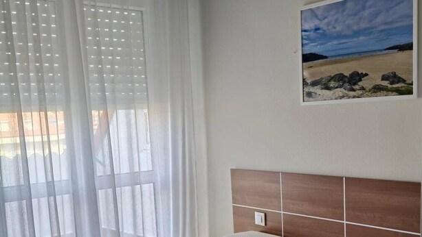 Habitación de hotel con cama, ventana con cortinas translúcidas, un cuadro de una playa y una colcha roja.