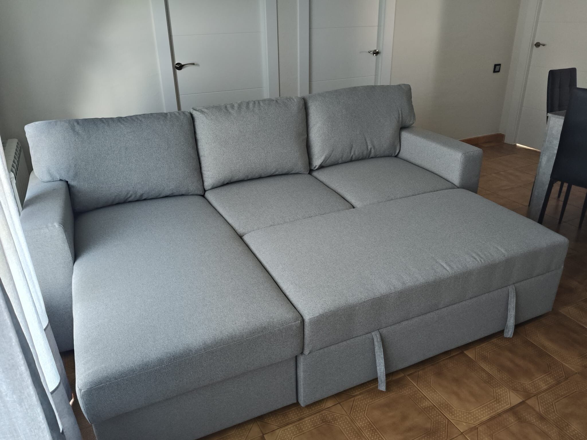 Un sofá seccional gris con reposapiés, una mesita y una silla en una habitación con suelo de baldosas y paredes blancas.