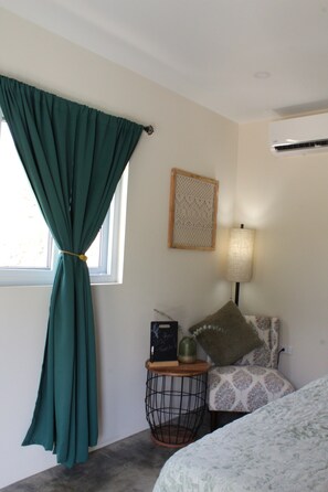 Un dormitorio con cama, mesita de noche, lámpara, ventana con cortinas y aire acondicionado.