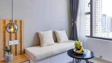 A modern living room with a sofa, a small round table with fruits, a hanging light, and a view of buildings through a window.
