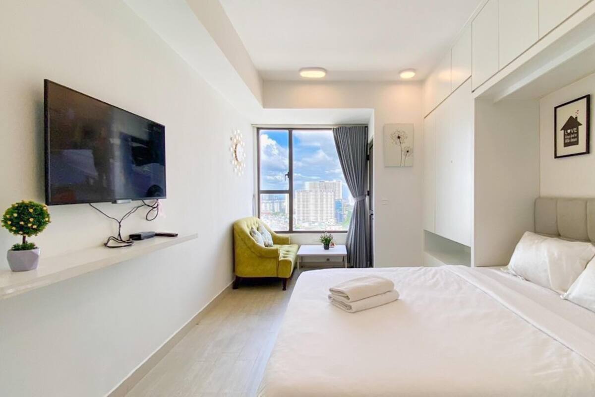 Ein modernes Hotelzimmer mit einem großen Bett, einem an der Wand befestigten Fernseher, einem kleinen Tisch mit Stuhl und einem Fenster mit Blick auf Gebäude.
