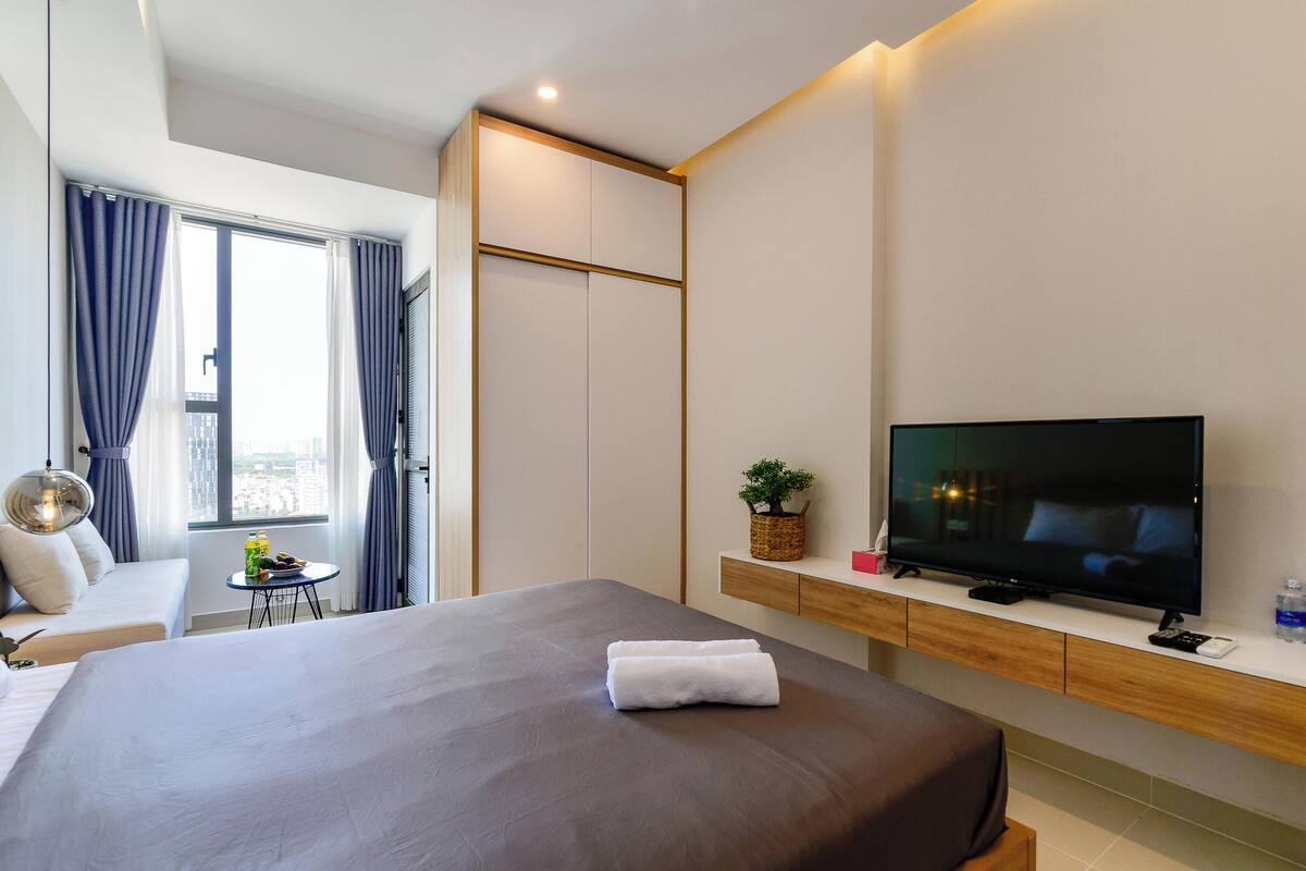 Ein modernes Hotelzimmer mit einem großen Bett, einem Fernseher, einem kleinen Tisch mit Pflanze und Blick auf die Stadt durch das Fenster.