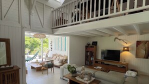 En rummelig stue med højt til loftet, en balkon, et fjernsyn og et stort vindue med udsigt over grønne områder.