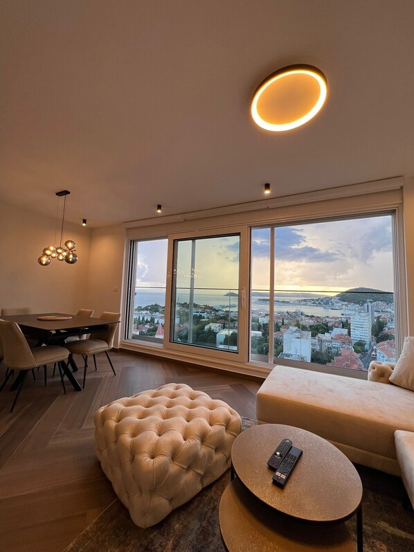A spacious living room with a large window offering a city view, a tufted ottoman, a round coffee table with a remote, and a dining area with a table and chairs.