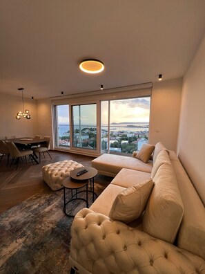 A modern living room with a large window, a sofa, a dining table, and a chandelier.