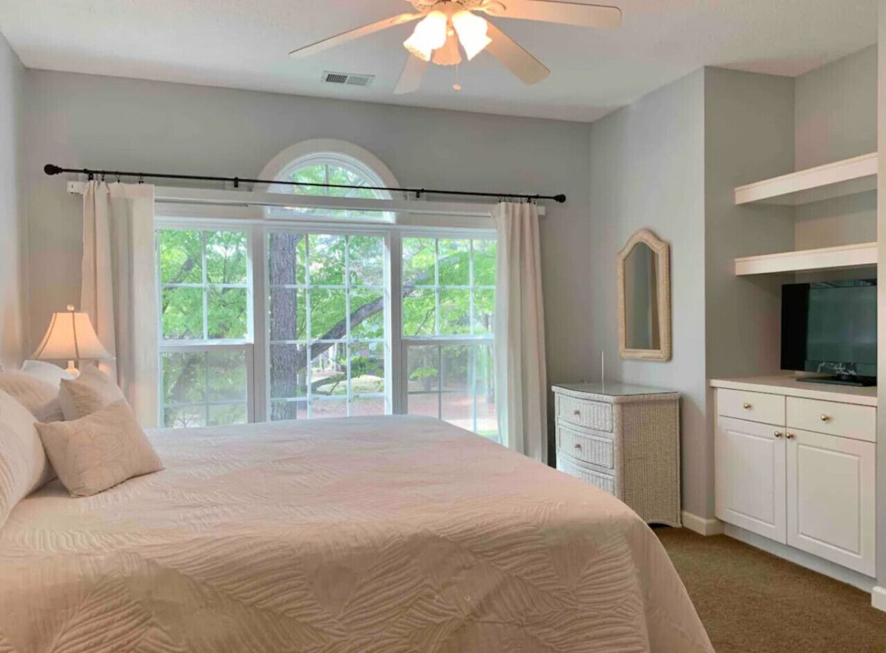 A bedroom with a large bed, a ceiling fan, a window with curtains, a dresser, a television, and a mirror.