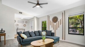 A modern living room with a dark blue sectional sofa, a wooden coffee table, and a large window with a view of a street and greenery.