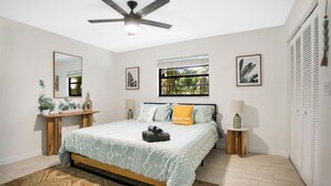 A bedroom with a bed, a ceiling fan, a window with a view of greenery, a bedside table with a lamp, and a mirror on the wall.