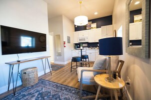 A modern living room with a flat-screen TV, a dining area with a table and chairs, a kitchen with white cabinets, and a large mirror.