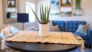 Et rundt bord med en vævet dug, en hvid potte med en aloe plante, en blå sofa og billeder i rammer på væggen.