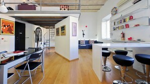 Eine moderne Loft-Wohnung mit einer Schlafgalerie, einem Essbereich mit Tisch und Stühlen, einer Küchenzeile mit Barhocker und einem Wohnzimmer mit Sofa und Zierkissen.