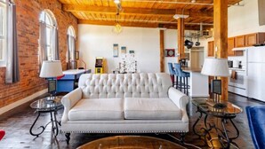 A spacious living area with a tufted sofa, two side tables with lamps, a wooden ceiling, and a brick wall. - Fun Loft with Arcade Games and River Views (Detroit)