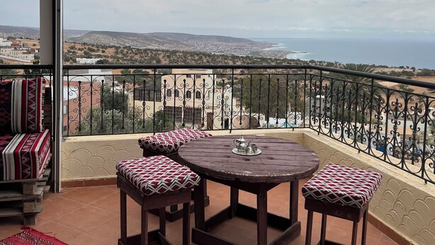 A balcony with a round table and four cushioned chairs, a view of a coastal landscape, and a railing.