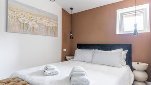 A neatly made bed with white linens and two folded towels, a tufted brown headboard, a wall-mounted painting of flowers, a black pendant light, and a bedside table with a vase. - Delightful apartment in Trastevere on two levels MauveSweetRhome 3 (Roma)