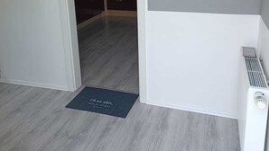 Een kamer met een grijze houten vloer, een radiator en een deur naar een keuken.