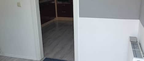 A room with a grey wooden floor, a radiator, and a door leading to a kitchen area.