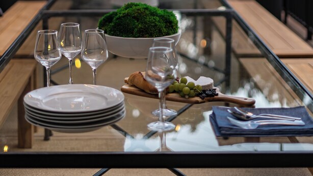 Eettafel buiten, met een glazen blad, omringd door stoelen, een schaal met groen en een bord met druiven.
