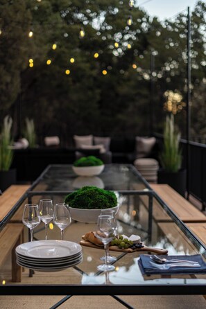 Une table à manger installée à l’extérieur, avec un plateau en verre, entourée de chaises, d’un saladier rempli de verdure et d’une assiette de raisins.