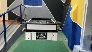 Une table de baby-foot dans une pièce dont le sol est vert, les rampes bleues et un mur orné de motifs géométriques.