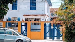 Un immeuble de deux étages avec une façade blanche et des volets bleus, une entrée fermée par un portail et une voiture garée devant.
