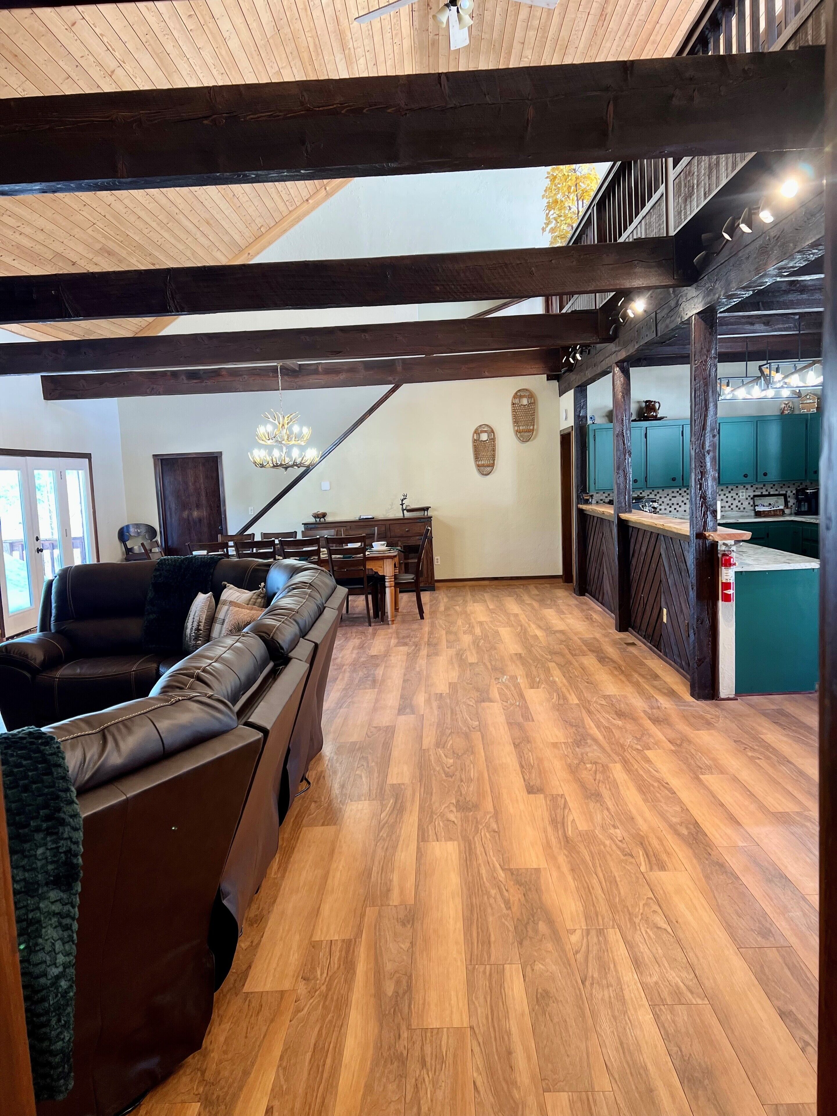 A spacious living area with a wooden ceiling, a dining table with chairs, and a kitchen area with teal cabinets.