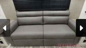 A grey sofa with a cushioned backrest and armrests, positioned in a room with a light-colored floor and a window with blinds.