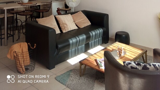 Un salon avec un canapé noir, une table basse en bois et un coin repas avec une cuisine en arrière-plan.