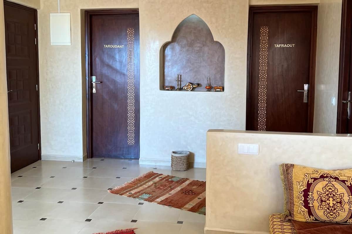 A hotel lobby with two wooden doors labeled "TAROUTANT," a decorative wall arch, a patterned rug, and a sofa with a patterned cushion.